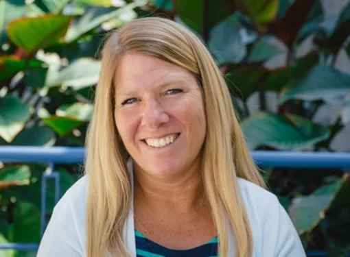 Michelle who has long blonde hair and is wearing a white cardigan and striped blue shirt. There are plants in the background.