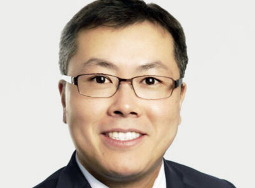 Ray Woo who has short dark hair, glasses and is wearing a dark suit and tie. He is against a light grey background.