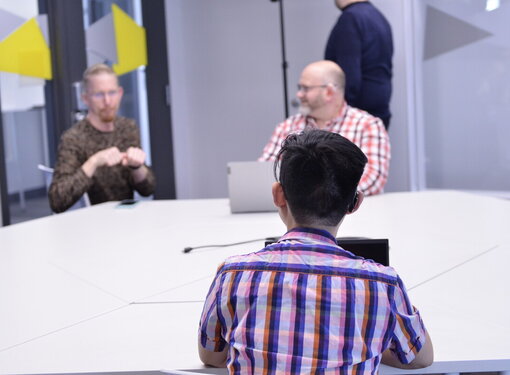 Back of the head of someone with short dark hair wearing a plaid shirt, has a laptop open and sitting at a table with two other people. One person is using American Sign Language