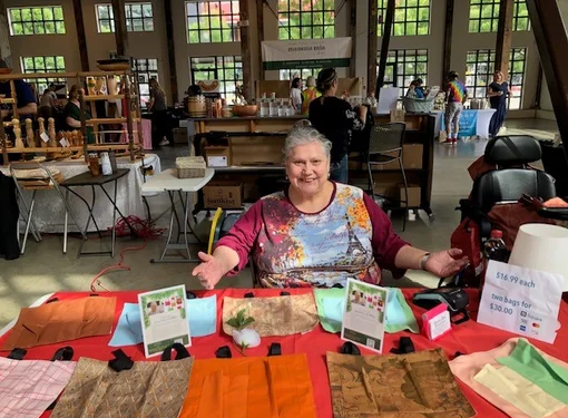 Julie Melanie, who has grey hair that is in a pony tail, sitting at a table with an orange table cloth that has her bags on display.