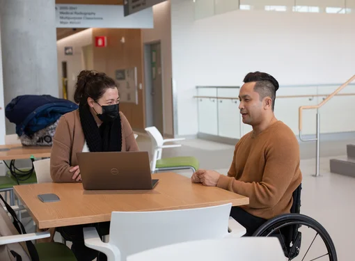 Two people working at a desk using a computer. One person is wearing a face mask and the other person is using a wheelchair.