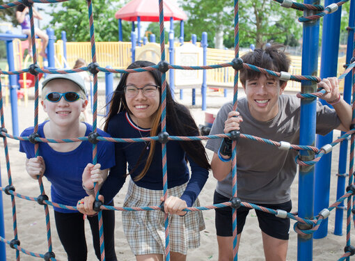 Three young people on a playground smiling and looking through a climbing rope. One youth has low vision and is wearing sunglasses.