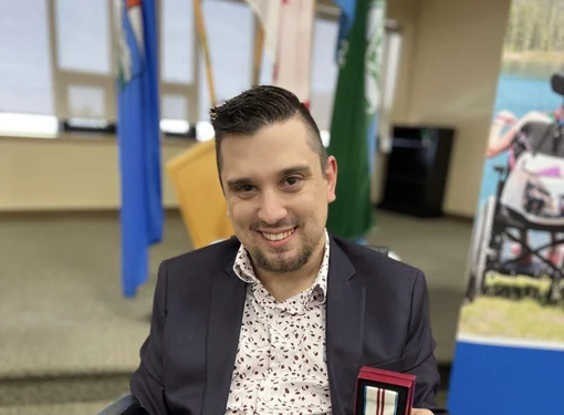 Brad Bartko, who has short brown hair and is wearing a suit jacket and a floral pattern button up shirt. He is using a wheelchair and holding up a medal.
