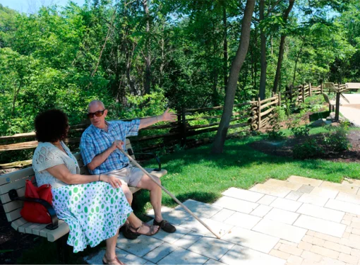 Two people sitting outside on a park bench talking. One person is using a white cane.