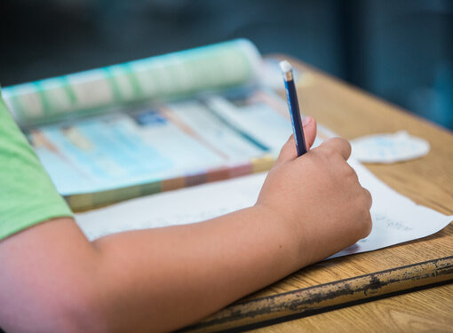 A child's arm writing on a piece of paper with a pencil. There is an open book on the desk.