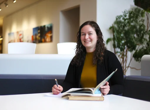 Amanda Motyer sitting at a table with an open book and pen. She has curly brown hair and is wearing a brown shirt and black blazer.