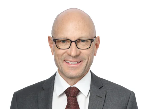 Man wearing glasses with a grey suit jacket, burgundy tie and white shirt.
