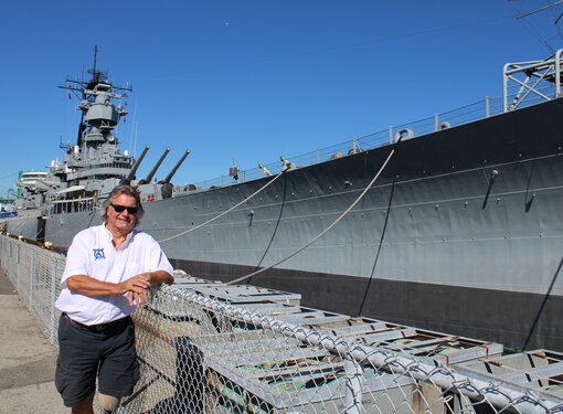 Colin McCarthy leaning on a chain link fence next to a large military ship. Colin has chin-length grey hair and is wearing a white shirt and black shorts.