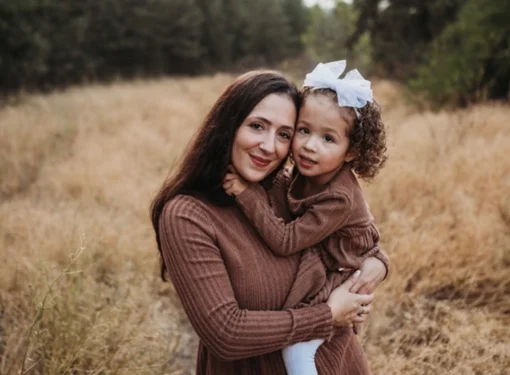 Katie holding her daughter Holland. They are wearing matching brown sweaters and are in front of a field.
