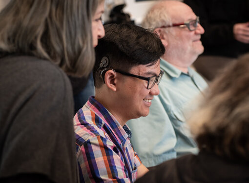 Person who is wearing glasses, has black hair and is wearing a cochlear implant smiling among a group of people.
