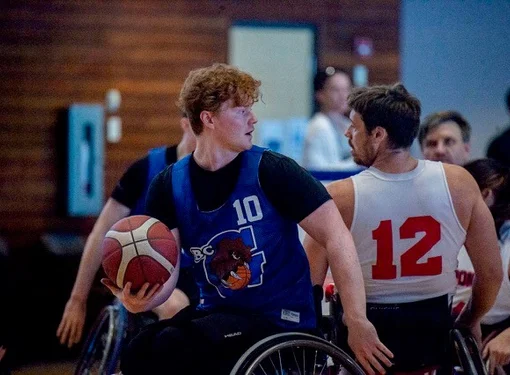 Cameron playing wheelchair basketball. He is holding the ball and looking back at an opponent. 