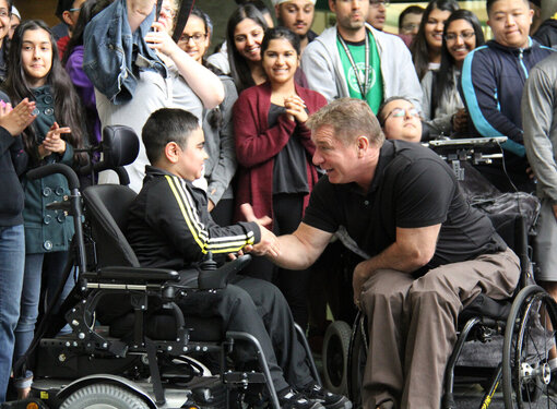 Rick Hansen shaking the hand of a young person who is using a wheelchair. A large crowd of students cheers behind them
