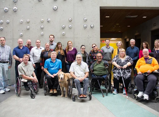 A group of people with different abilities smiling in front of a grey wall.