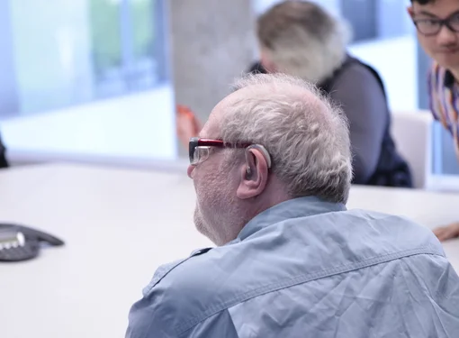AN elderly man who is wearing glasses and has a hearing aid. He is sitting at a table with others.