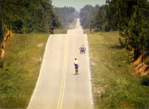 Rick Hansen during the Man In Motion World Tour. He is in the distance wheeling up a very large hill. A person on a bicycle rides behind him.