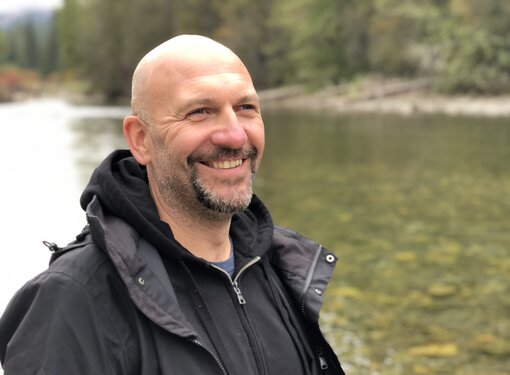 A man with a salt and pepper coloured beard by a river. He is wearing a black sweater and rain jacket and smiling.