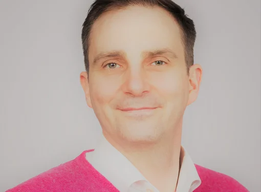 Headshot of a man with shirt dark brown hair and green eyes wearing a red shirt with a white collar underneath.