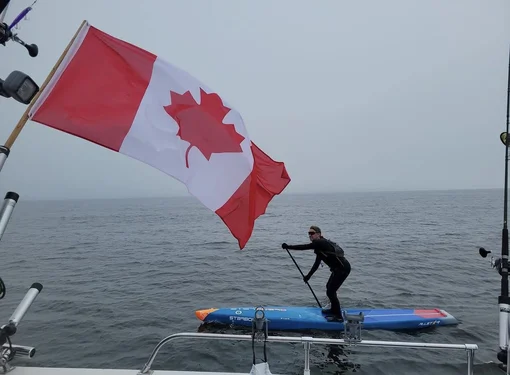 Mike Shoreman on a paddleboard in the lake. There is a large Canada flag in front of him