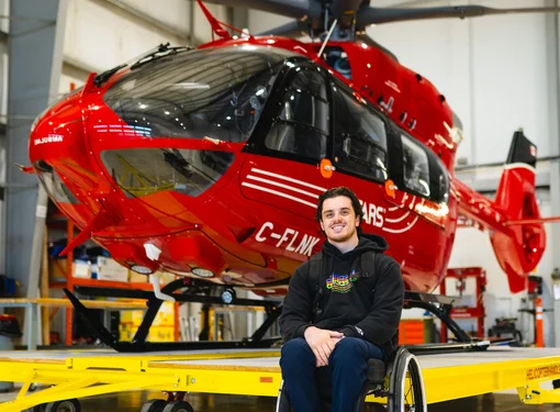 Ryan in front of a large red helicopter inside a garage. Ryan is using a wheelchair.