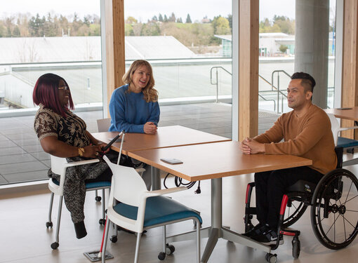 A woman with a white cane, a woman with a hearing aid and a man in a wheelchair sit around a table
