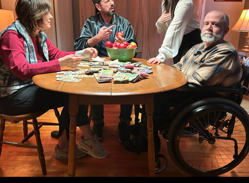 4 people argue around a table with wads of cash on it. One of them; a gentleman in a wheelchair stares at the camera