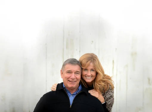 Rick and Amanda Hansen, smiling against a white backdrop, with Amanda hugging the Rick lovingly from behind.