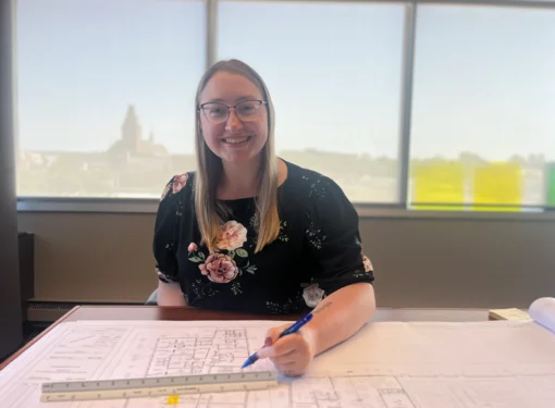 Cassie sitting at a table with architectural plans on it. She is wearing a black shirt with floral pattern on it. 