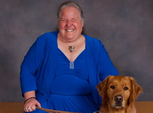 A woman who is blind smiles while seated, wearing elegant attire. A dog sits to her left. The background is dark and simple