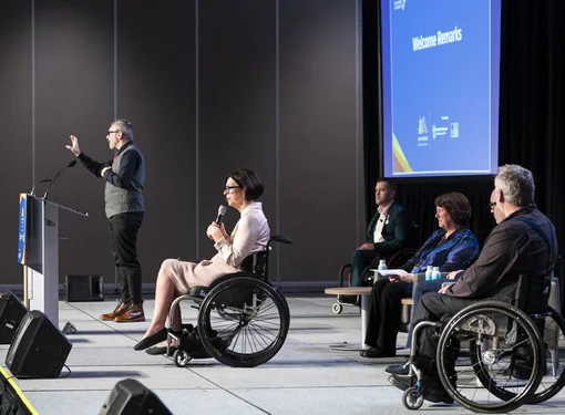 Five individuals are on stage, four of whom are seated, with three in wheelchairs. One person stands at a podium presenting, while another holds a microphone. In the background, a large blue screen displays various logos and branding elements.