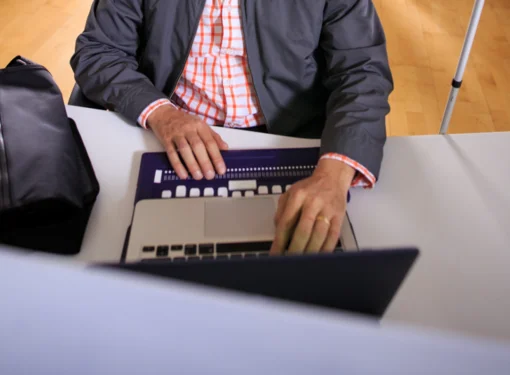 A man sits at a desk in an office, using an accessible Braille keyboard. He faces a computer monitor, with a white cane resting on the right side