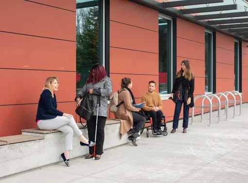 group of five adults outside a modern building. One person is sitting on a concrete bench, two people are standing and talking, one person is using a wheelchair, and another is standing nearby with a white cane