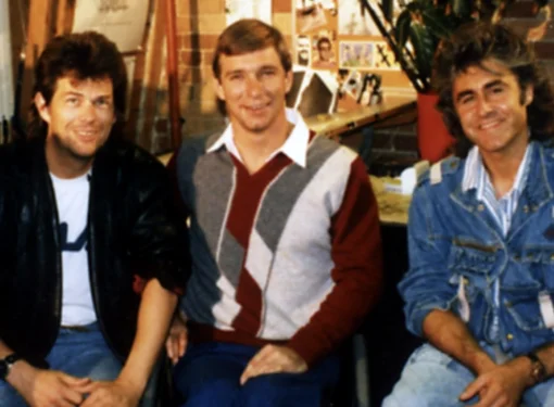 Three men sit together in a music studio, surrounded by desks, shelves, and papers pinned to the wall. They wear casual clothing typical of the late 1980s.