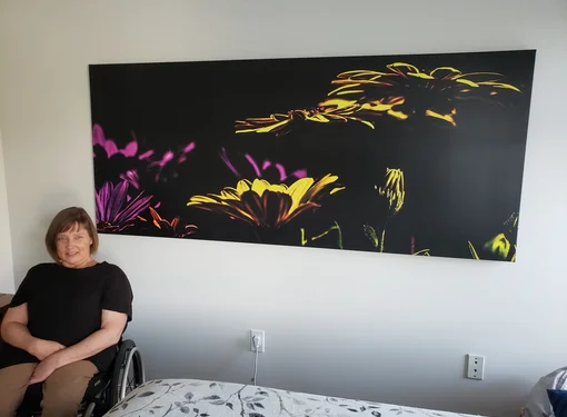 A woman (Marika) seated in a wheelchair in a bright room. A large wall art photo piece above the bed shows vivid yellow and purple flowers on a black background.