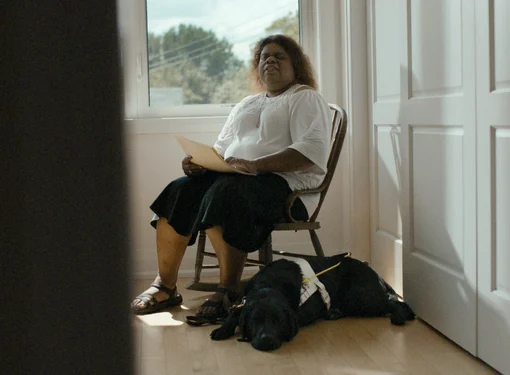 A person sits on a wooden chair in a bright room holding a document, with a guide dog lying on the floor beside them near a window.