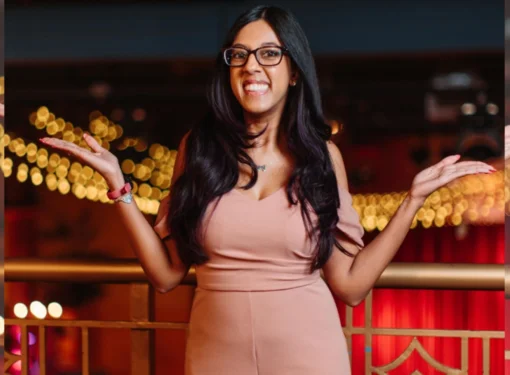 Woman with black long hair wearing glasses stands indoors near a railing, gesturing with both hands, with decorative lights and red drapery in the background.