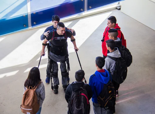 Person wearing a robotic exoskeleton stands using forearm crutches while a group of youth with backpacks gathers around and listens.