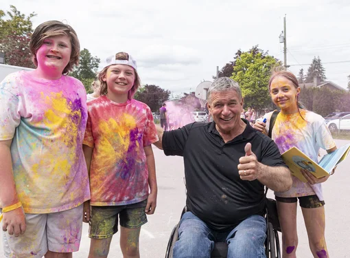 Three children covered in colorful powder stand outdoors beside Rick. One child holds an open book while the group poses on a school playground.