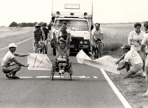 old photo of Rick wheeling while crossing a marked line on a rural road with both arms raised. Supporters hold signs on either side of the road, while cyclists and a support vehicle follow behind.
