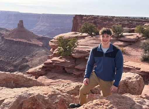 Young man with short, dark curly hair wearing a blue pullover and tan pants, standing among red rock formations overlooking a vast desert canyon with layered cliffs and scattered shrubs in the background.