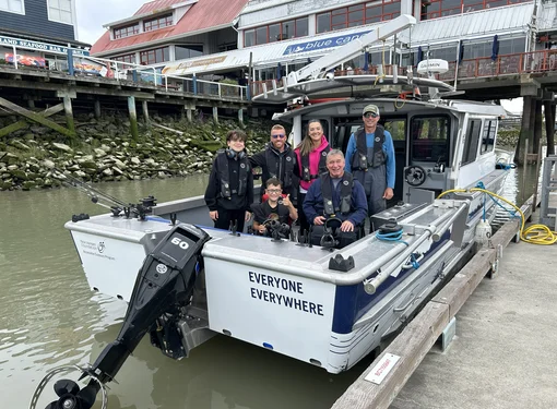 A group of five people aboard an accessible fishing boat docked at a marina. A man and a boy are using wheelchairs and are positioned near the centre of the group.