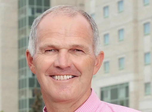 Middle aged man with grey short hair, blue eyes, smiling, wearing a pink button up shirt with tie. 
