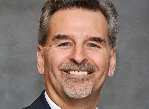 Middle aged man, grey hair, smiling, wearing a grey suit. 