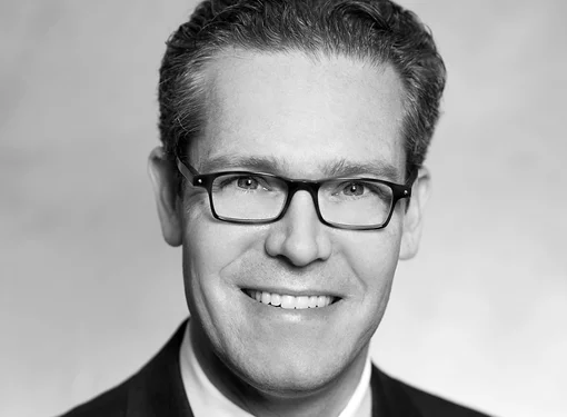 Middle aged man, short hair, glasses, wearing black suit with tie. 