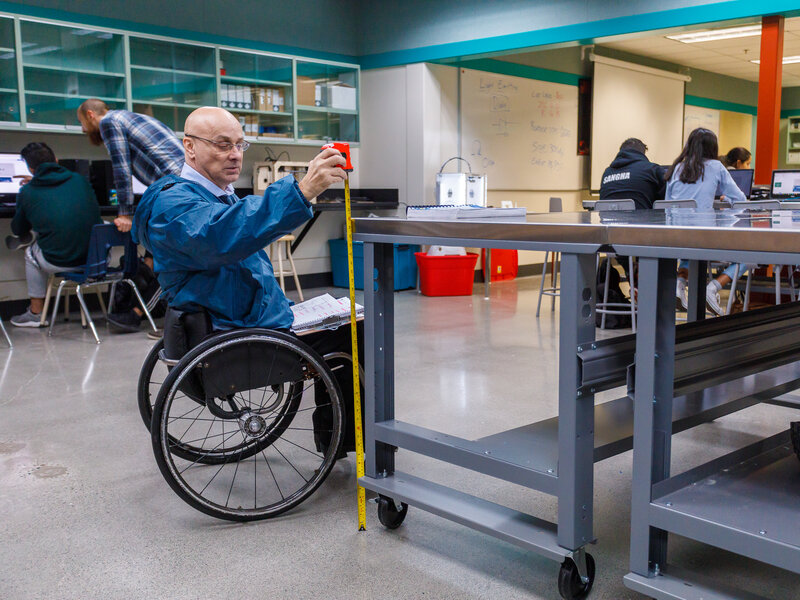 Man using wheelchair holds a measuring tape to measure a table. 
