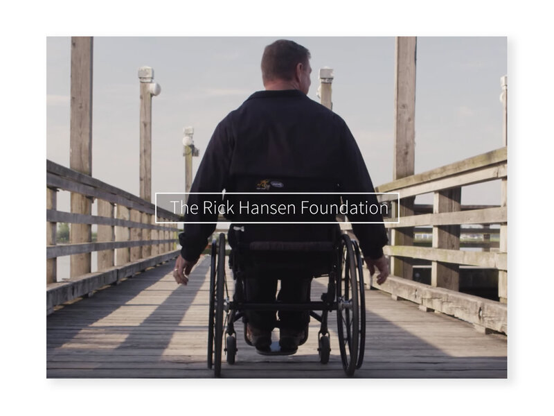 Screenshot from "Changing the Conversation About Disability" video of Rick Hansen's back as he wheels forward on a wooden bridge. The words "The Rick Hansen Foundation" are overlayed in the middle.
