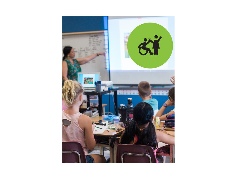 Backs of students sitting at desks, looking at teacher who is at the white board pointing to a projected screen. Small green circle icon with graphic of person in a wheelchair high-fiving a person that is standing.