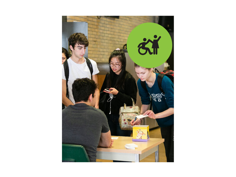 Group of teenagers standing around a desk. Small green circle icon with graphic of person in a wheelchair high-fiving a person that is standing.