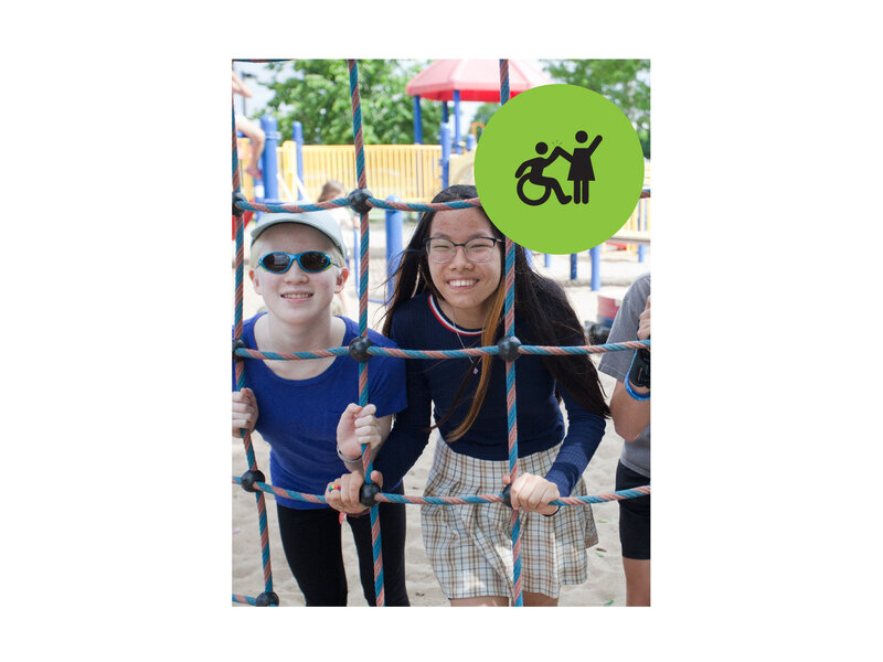 Two teenage girls standing behind and holding onto a net climbing wall at a playground. Small green circle icon with graphic of person in a wheelchair high-fiving a person that is standing.