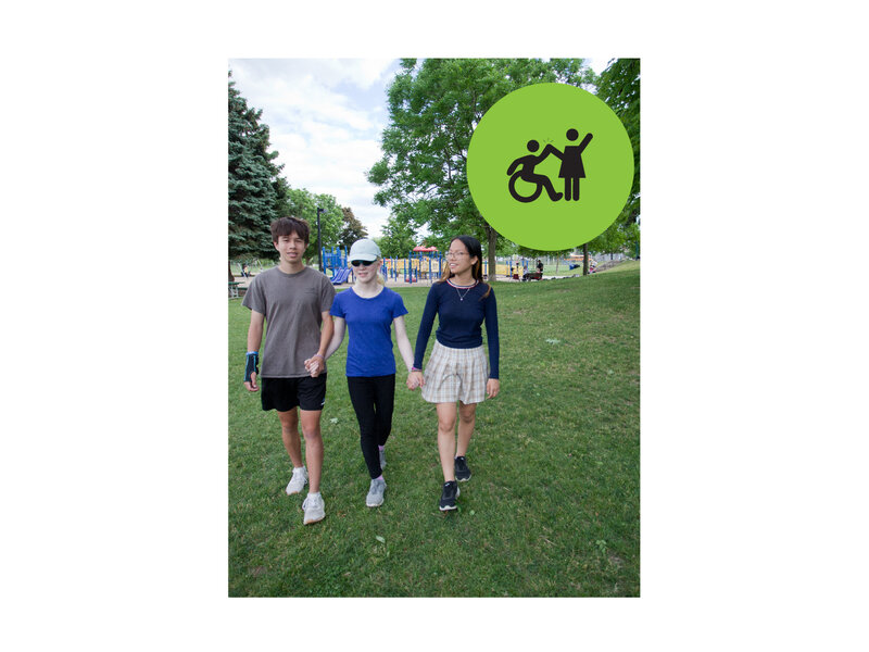 Group of 3 teenagers walking in a park, hand in hand. Small green circle icon with graphic of person in a wheelchair high-fiving a person that is standing.