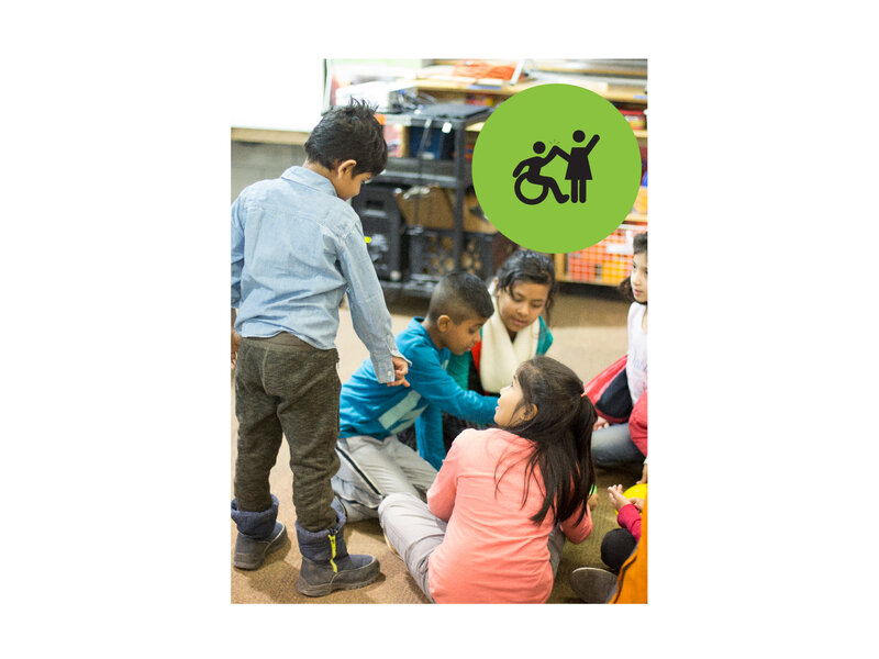 Boy going up to a group of children that are sitting and playing with blocks. Small green circle icon with graphic of person in a wheelchair high-fiving a person that is standing.